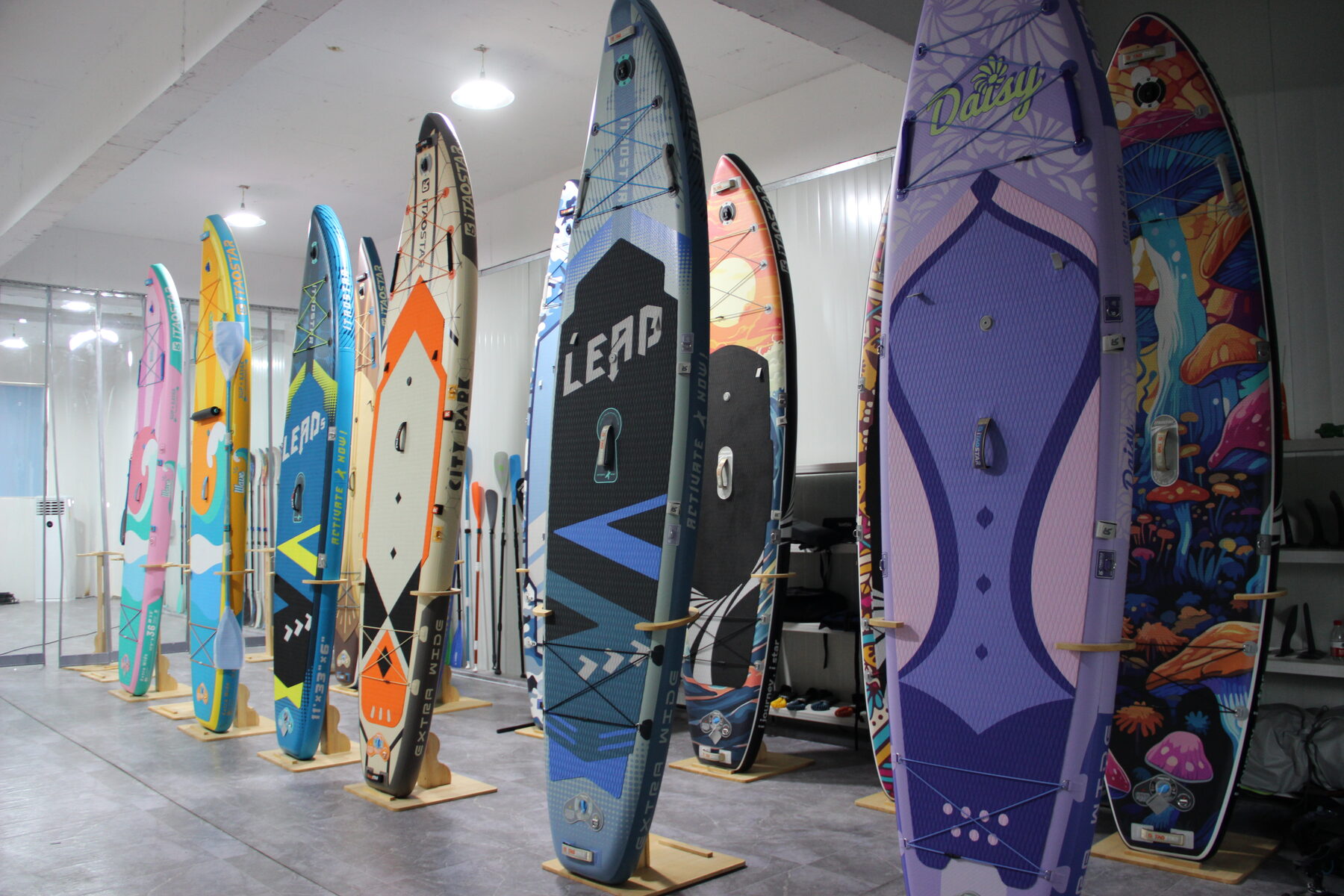 A row of finished paddle boards standing on end, in a range of colours and graphic designs.